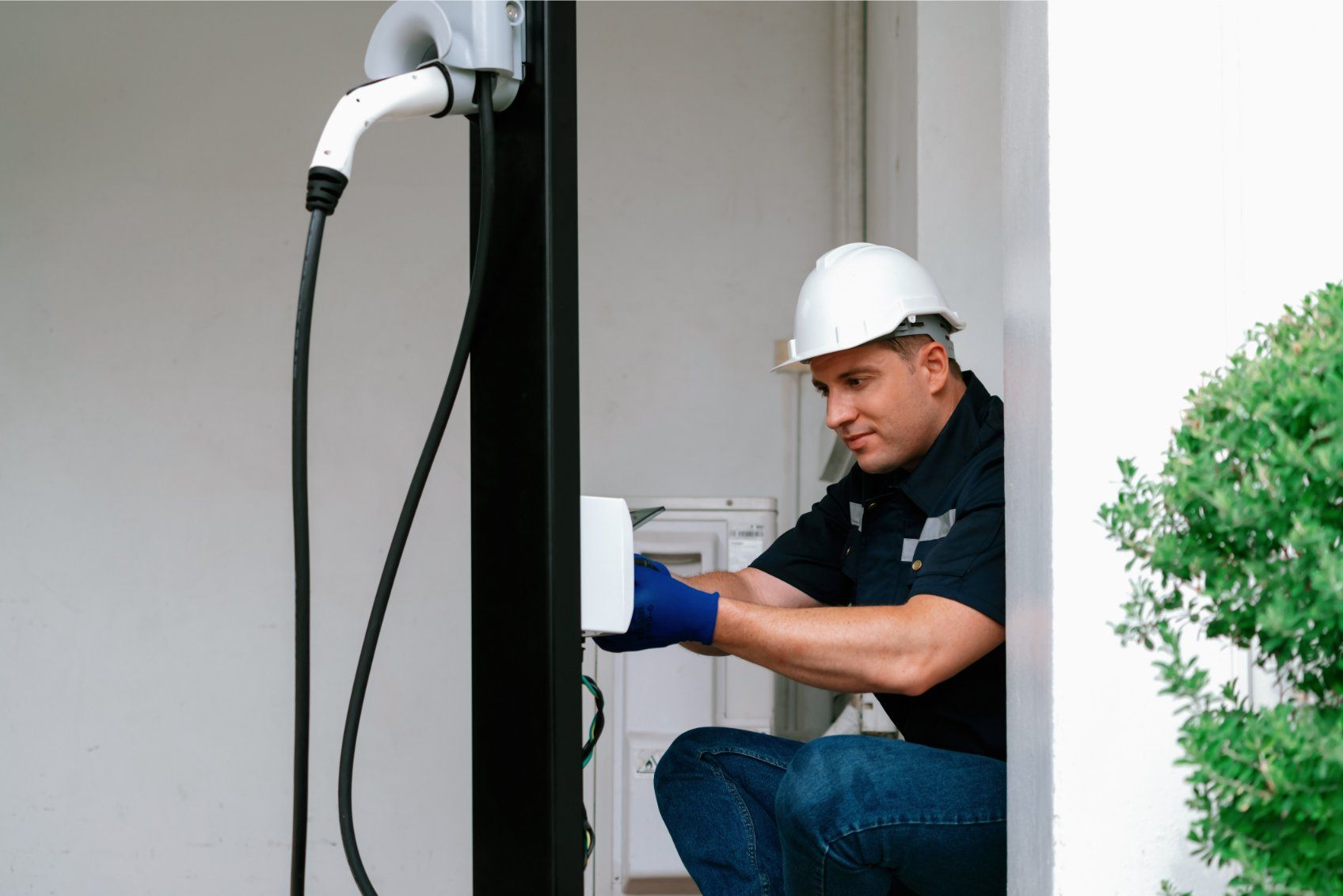Electrician installing an EV charger on a black pole; white hard hat, blue jeans.