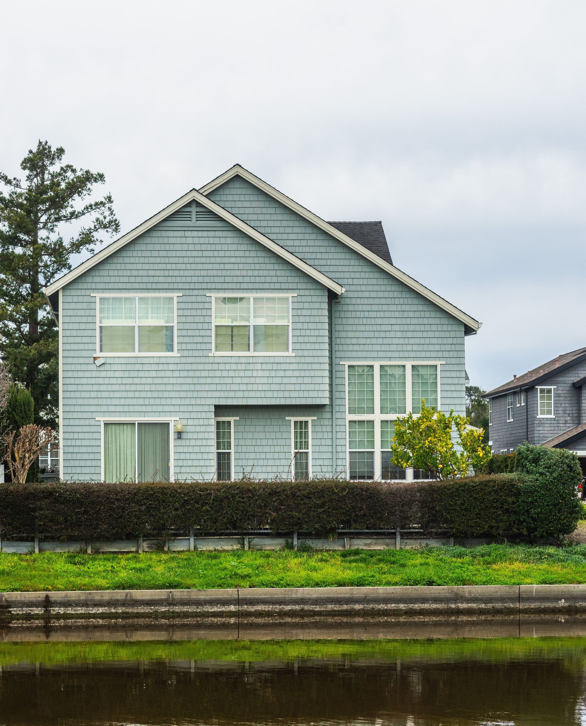 Light blue house with white trim behind a hedge beside a calm canal.