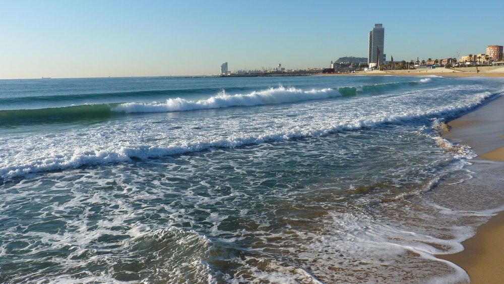 Una playa con olas rompiendo en la arena y una ciudad al fondo.