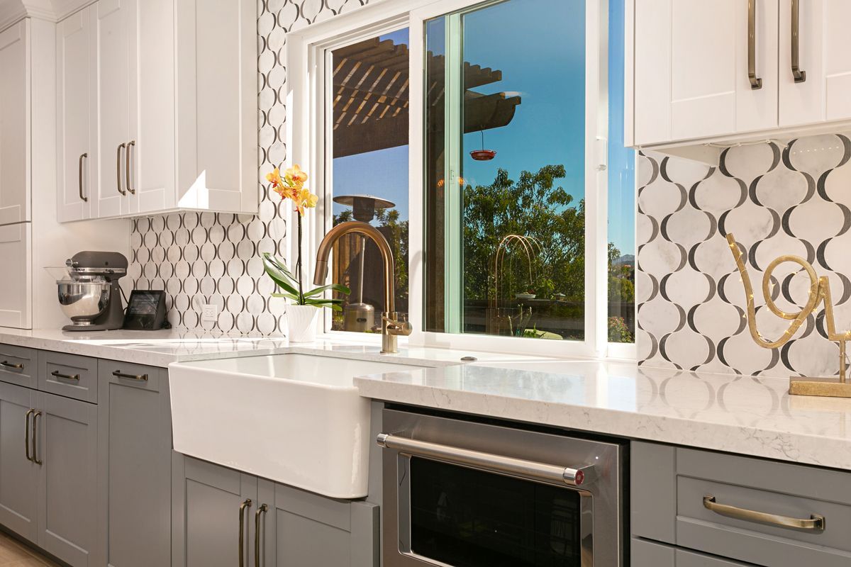 A kitchen with gray cabinets , white cabinets , stainless steel appliances and a large window.