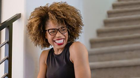 Woman with curly hair and glasses smiles broadly near a staircase.