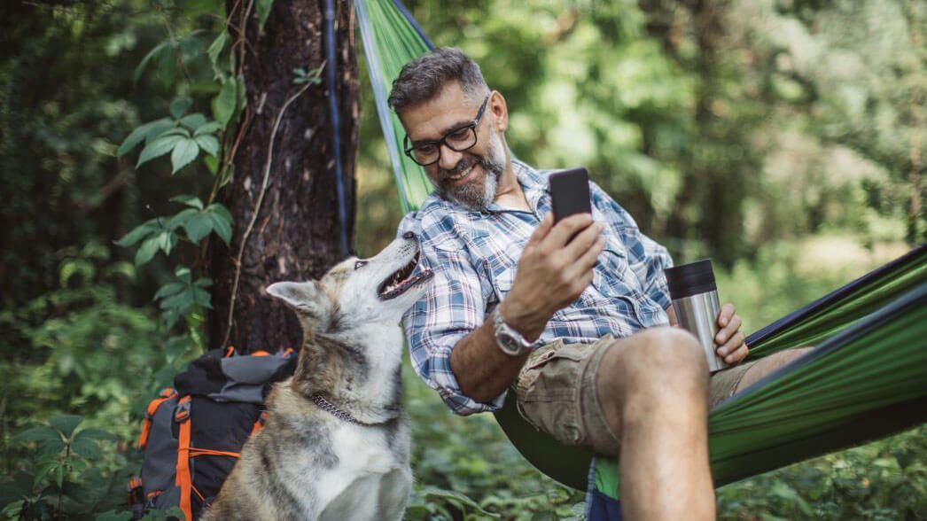 Man in hammock with dog, using phone. Forest setting. Man smiles, dog looks up.