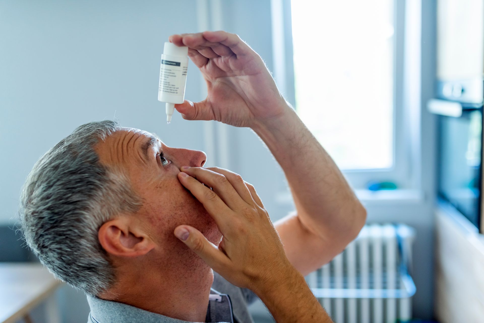 Man administering eye drops; indoors, looking up, holding bottle.
