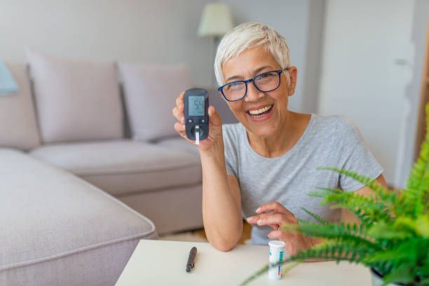 Woman with short gray hair smiles, holding a blood glucose meter, sitting at table with supplies, indoor setting.