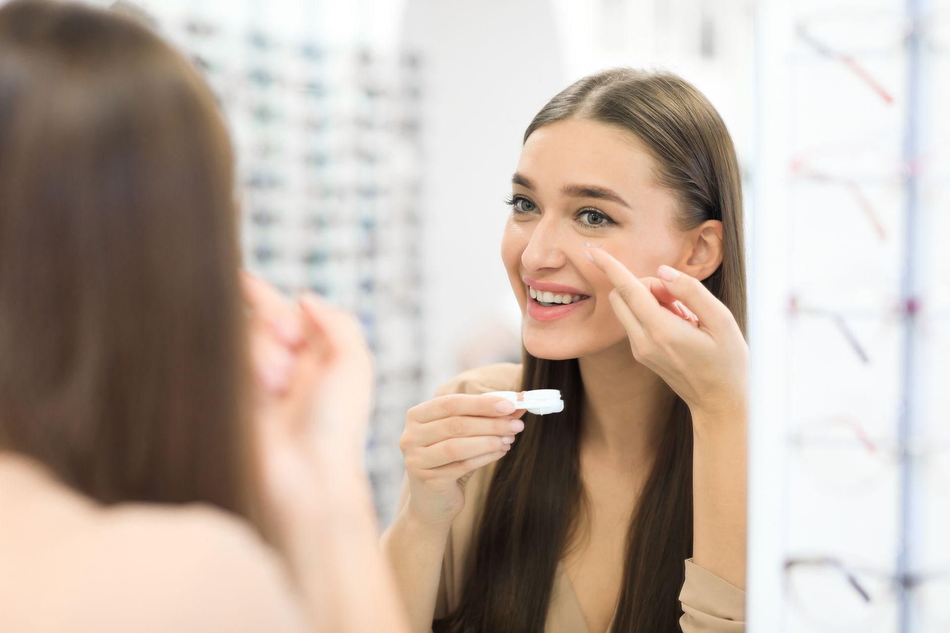 Woman inserts contact lens in mirror, smiling; eyewear store in background.