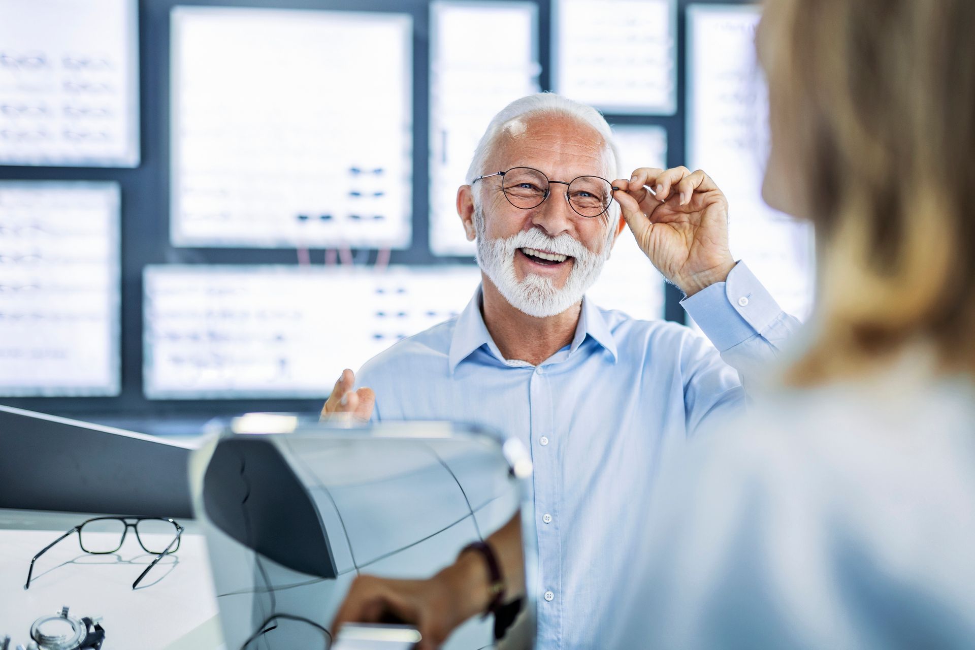 Man smiling, adjusting glasses, in an eyeglasses store, interacting with another person.