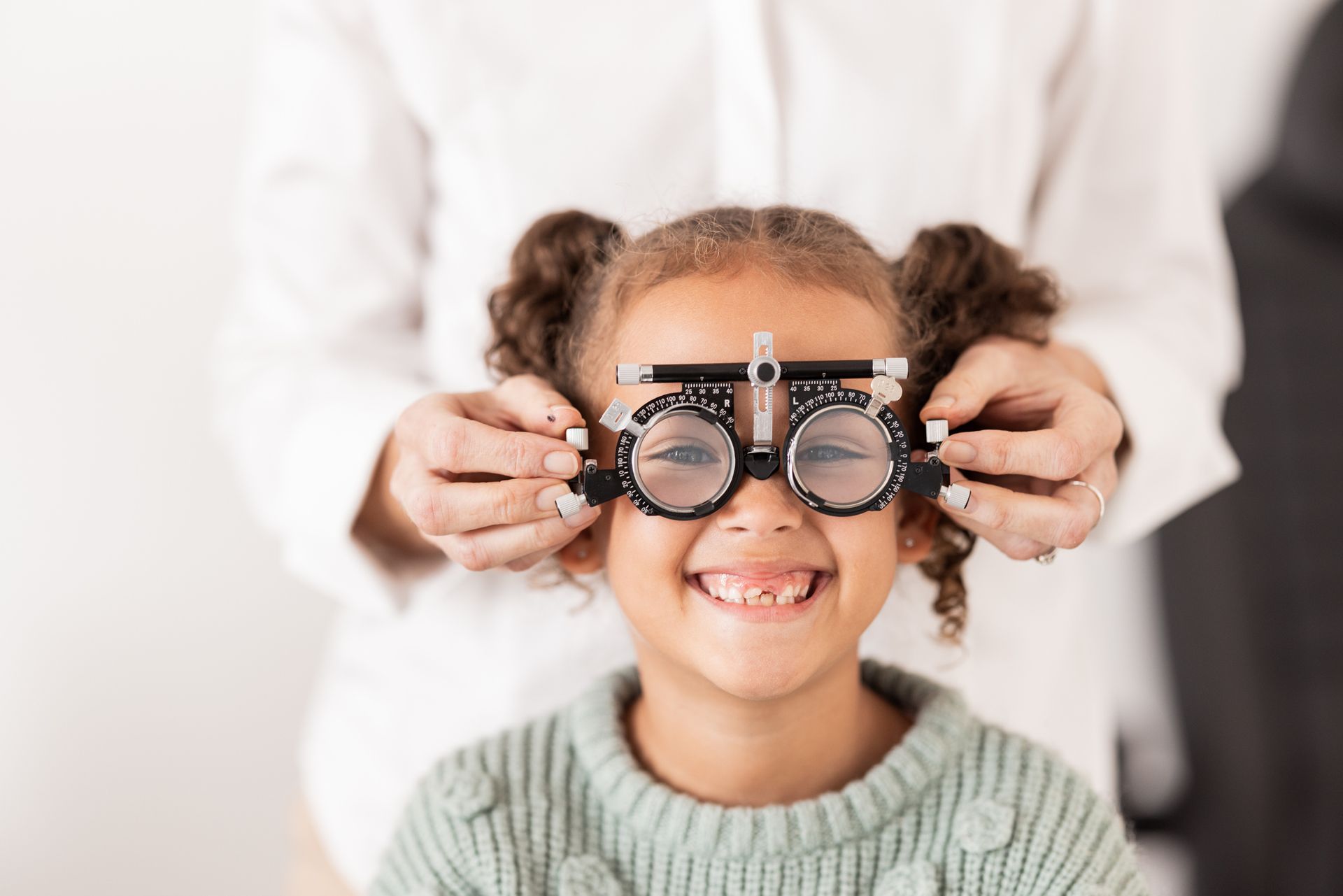 Girl wearing eye exam device, smiling, in an optometry office. Hands are adjusting the device.