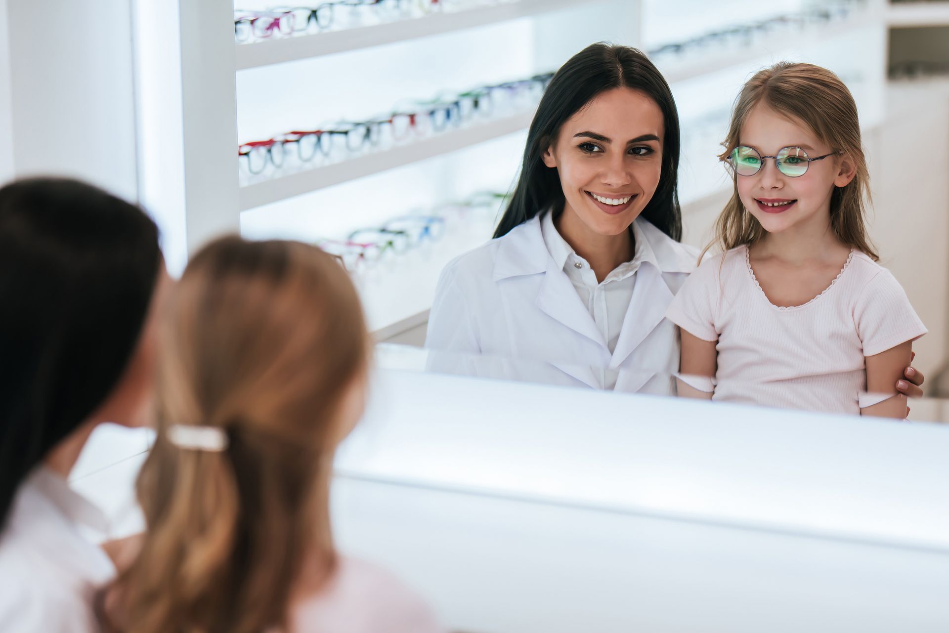 Optometrist with girl in glasses looking in a mirror in an eyewear store. They are smiling.