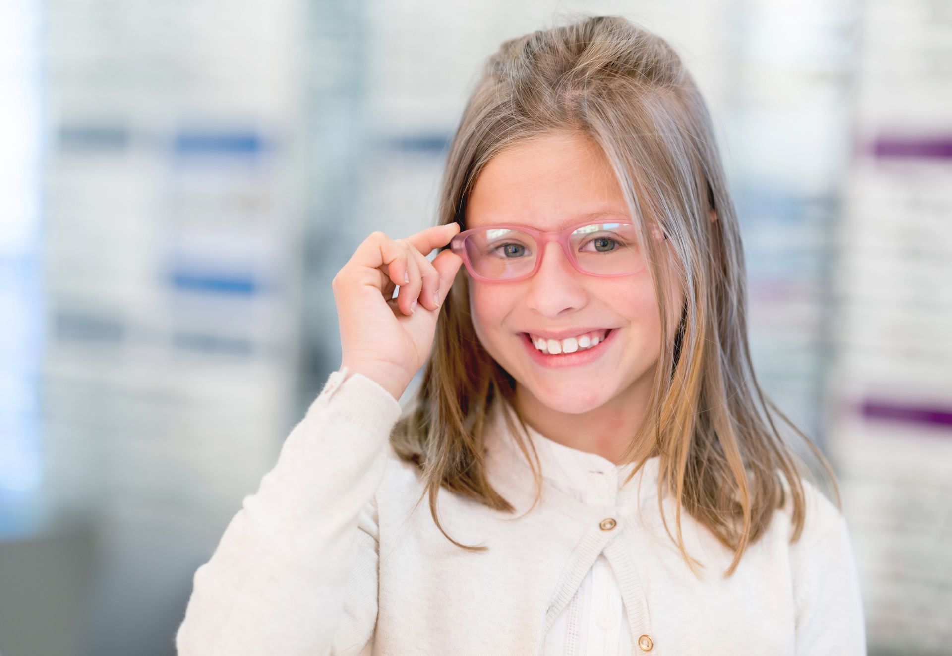 Girl smiling, adjusting pink glasses, in an eyeglasses store.