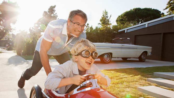 Man pushing a child in a red toy race car outdoors on a sunny day.