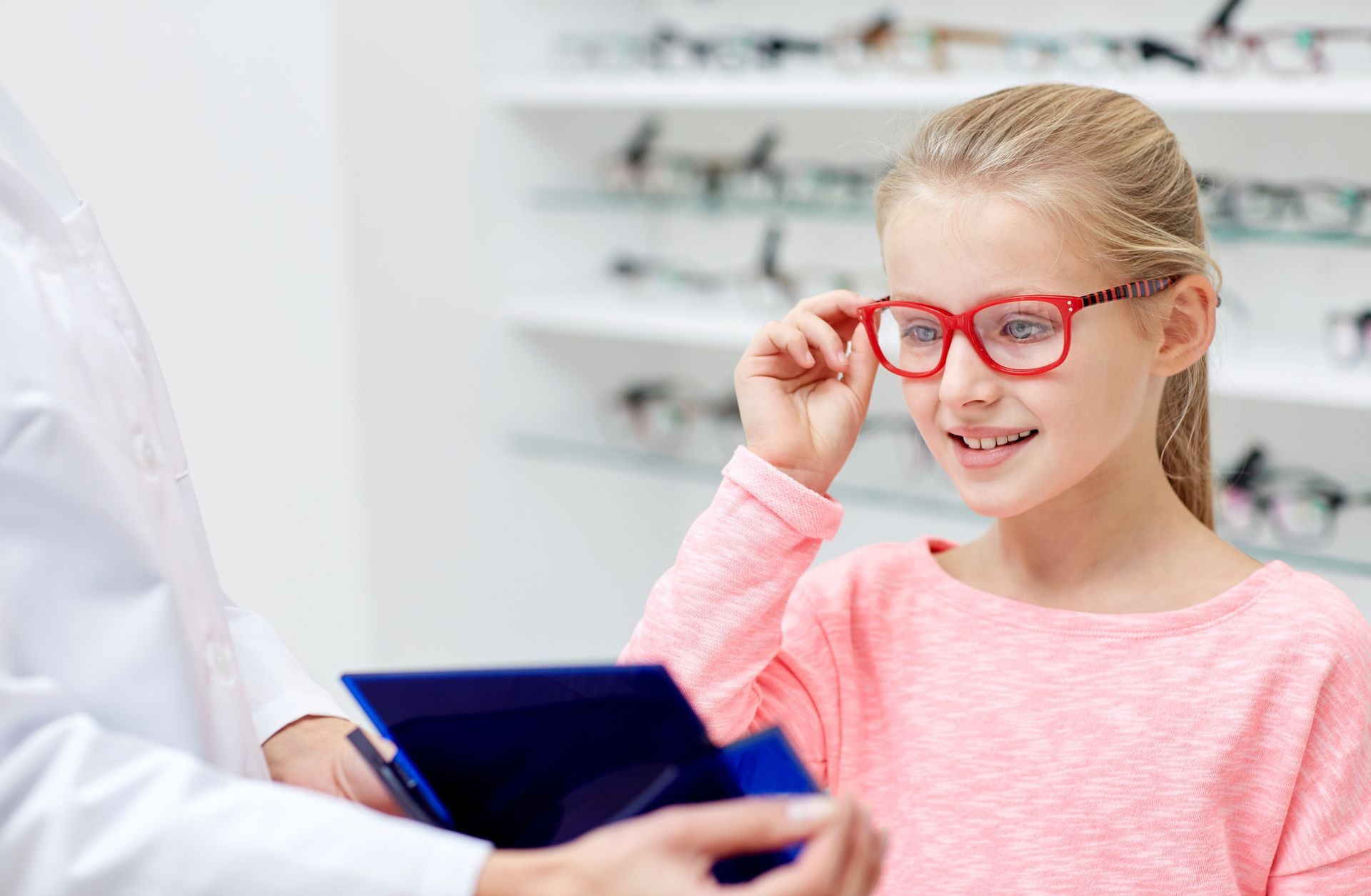Girl trying on red eyeglasses in an optician's office. An assistant holds the case.