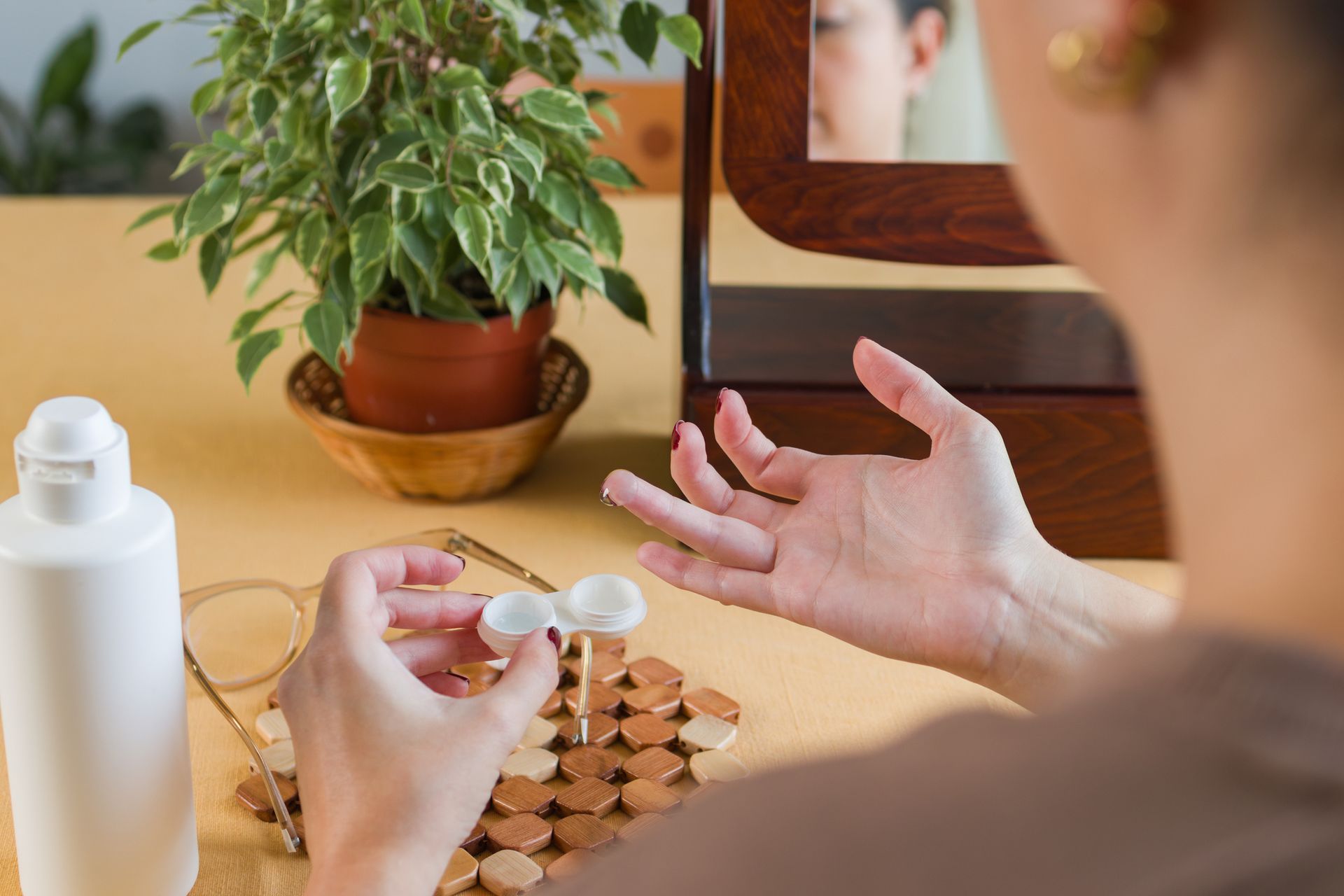 Woman preparing to insert contact lenses, holding one in her hand near a mirror, glasses, and a bottle of solution.