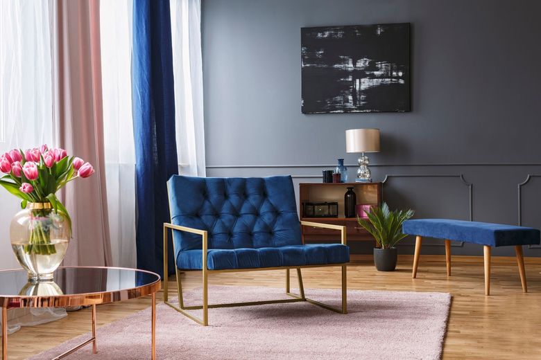 Blue tufted armchair with gold frame, pink rug, tulips, and art against a gray wall.