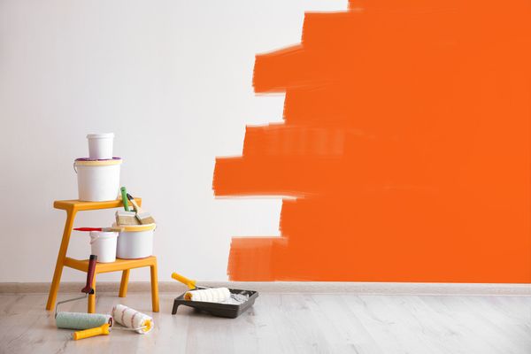 Orange paint partially covers a white wall. Paint cans, rollers, and a tray sit on a step stool and floor.