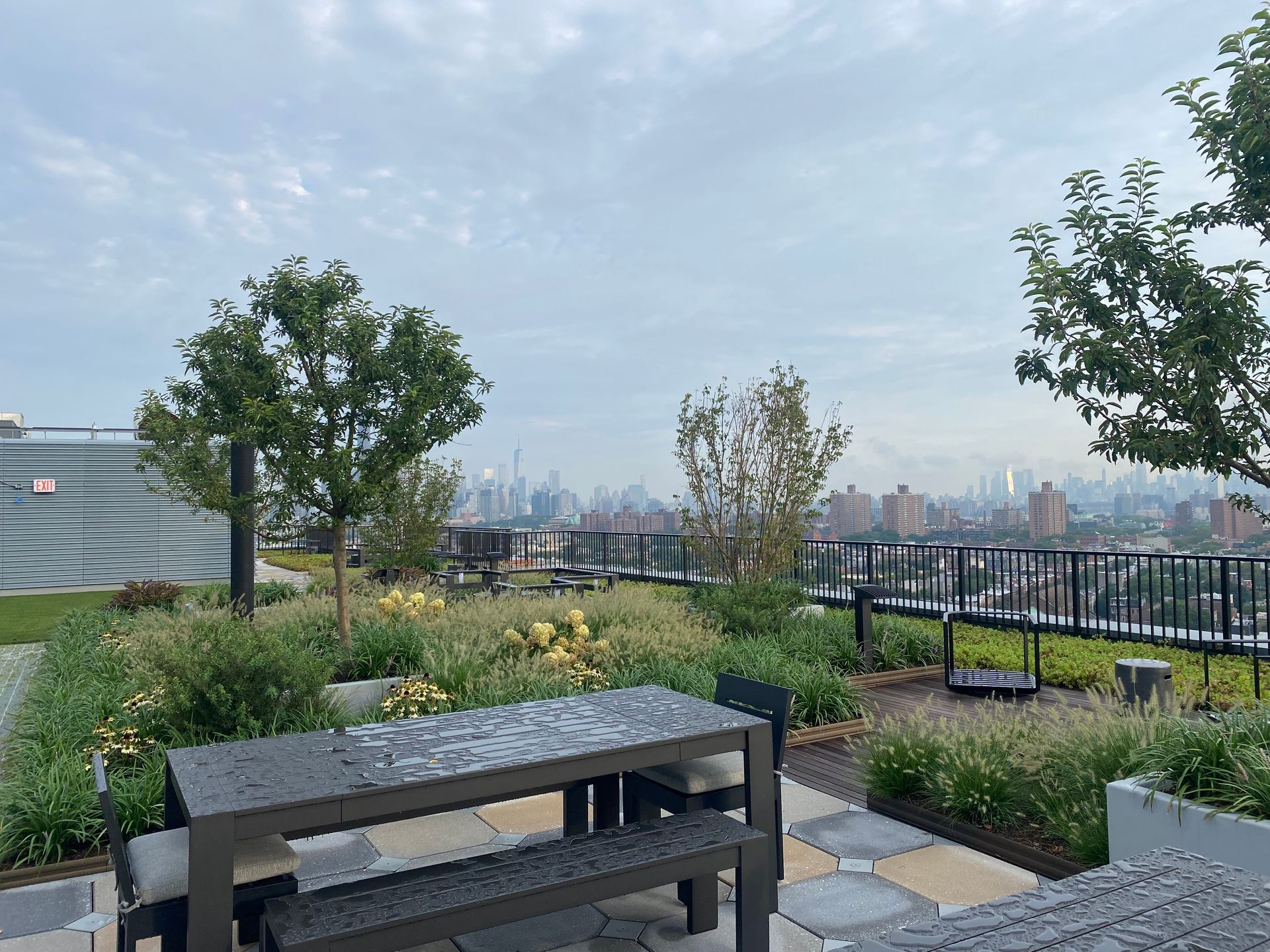 Rooftop garden with table, benches, plants, trees, and a city skyline under an overcast sky.