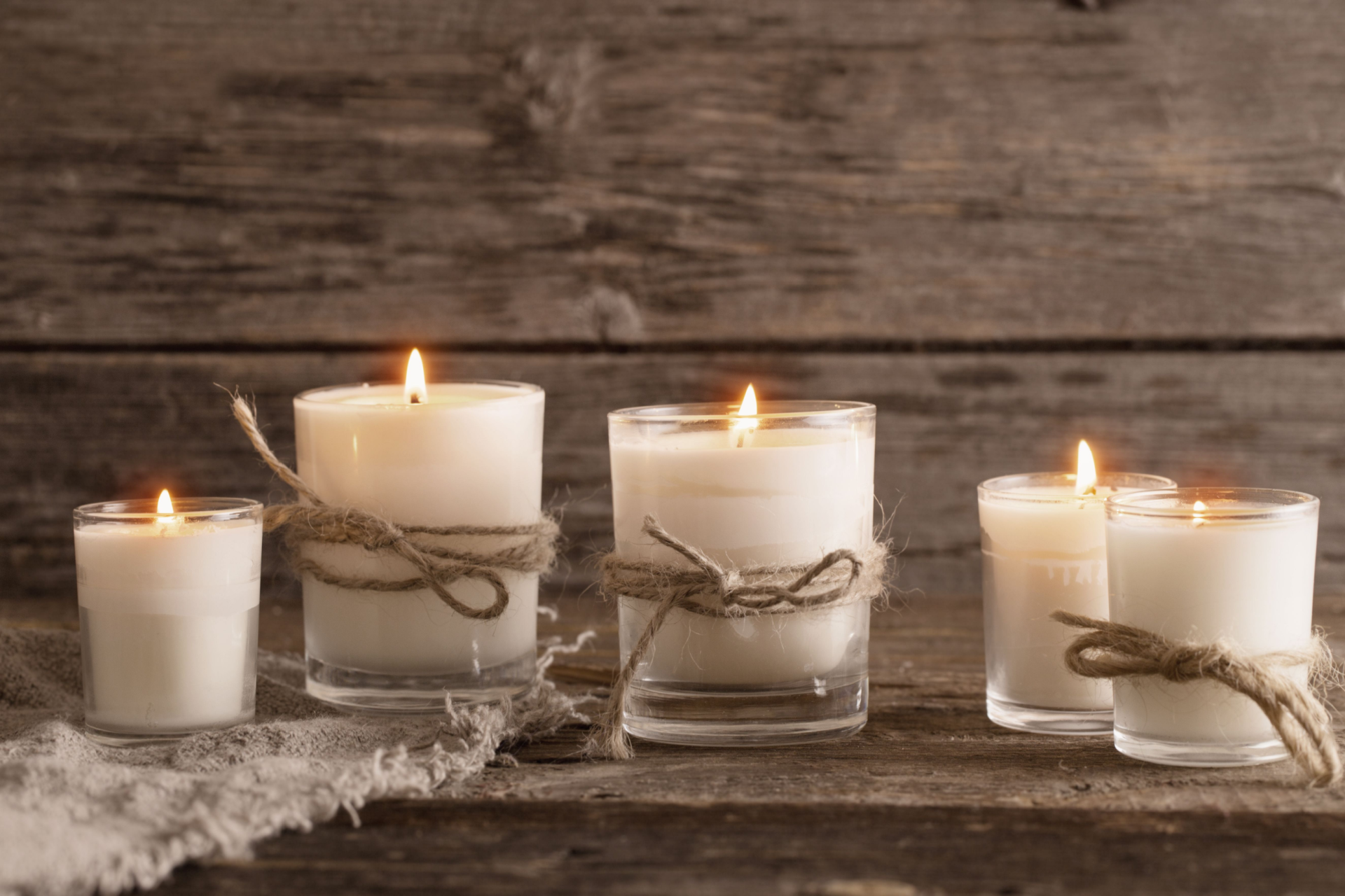 Five white candles in glass jars, tied with twine, sit on a wooden surface against a rustic wood background.