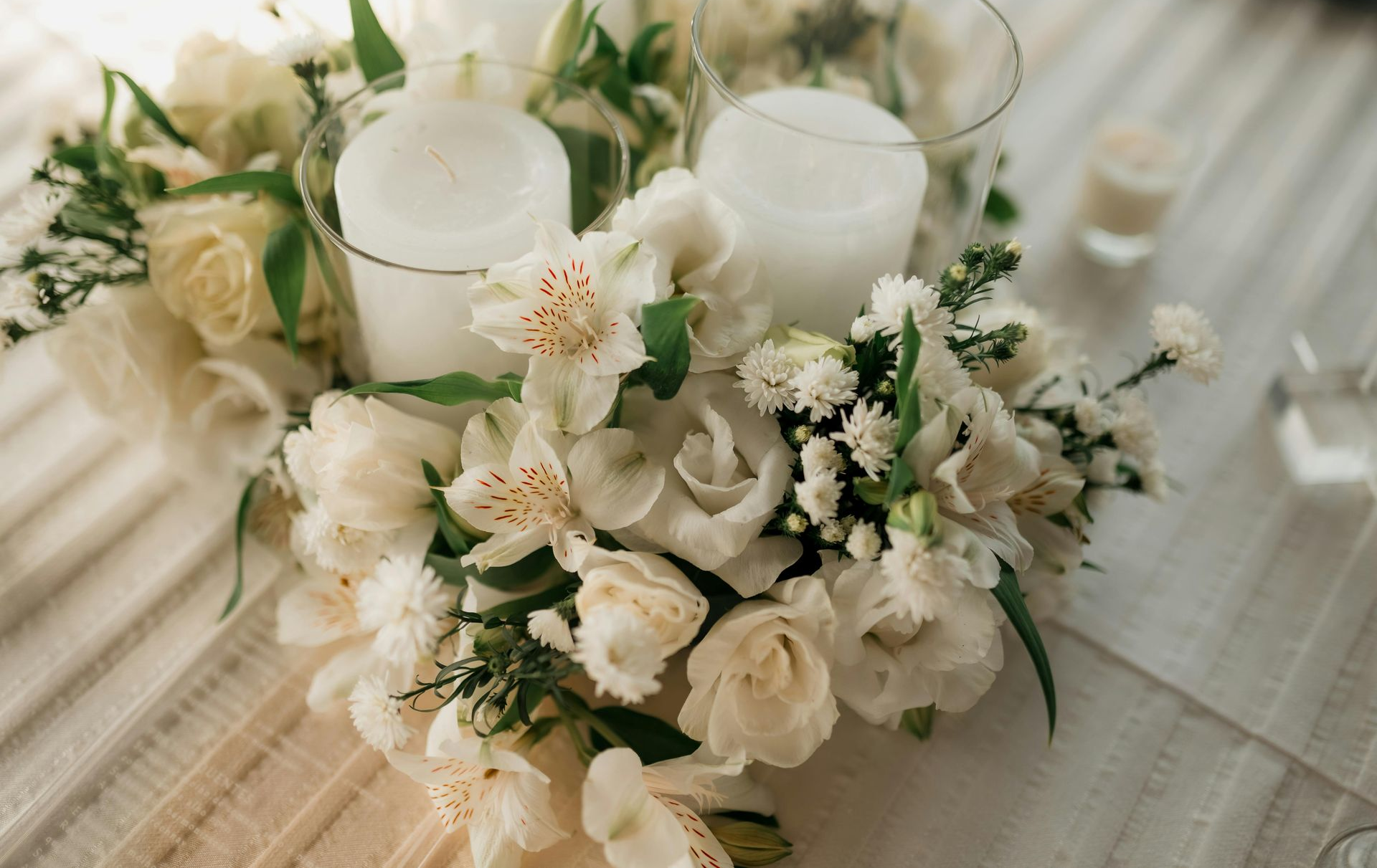Two white pillar candles in glass holders are surrounded by a lush arrangement of white roses and alstroemeria flowers.