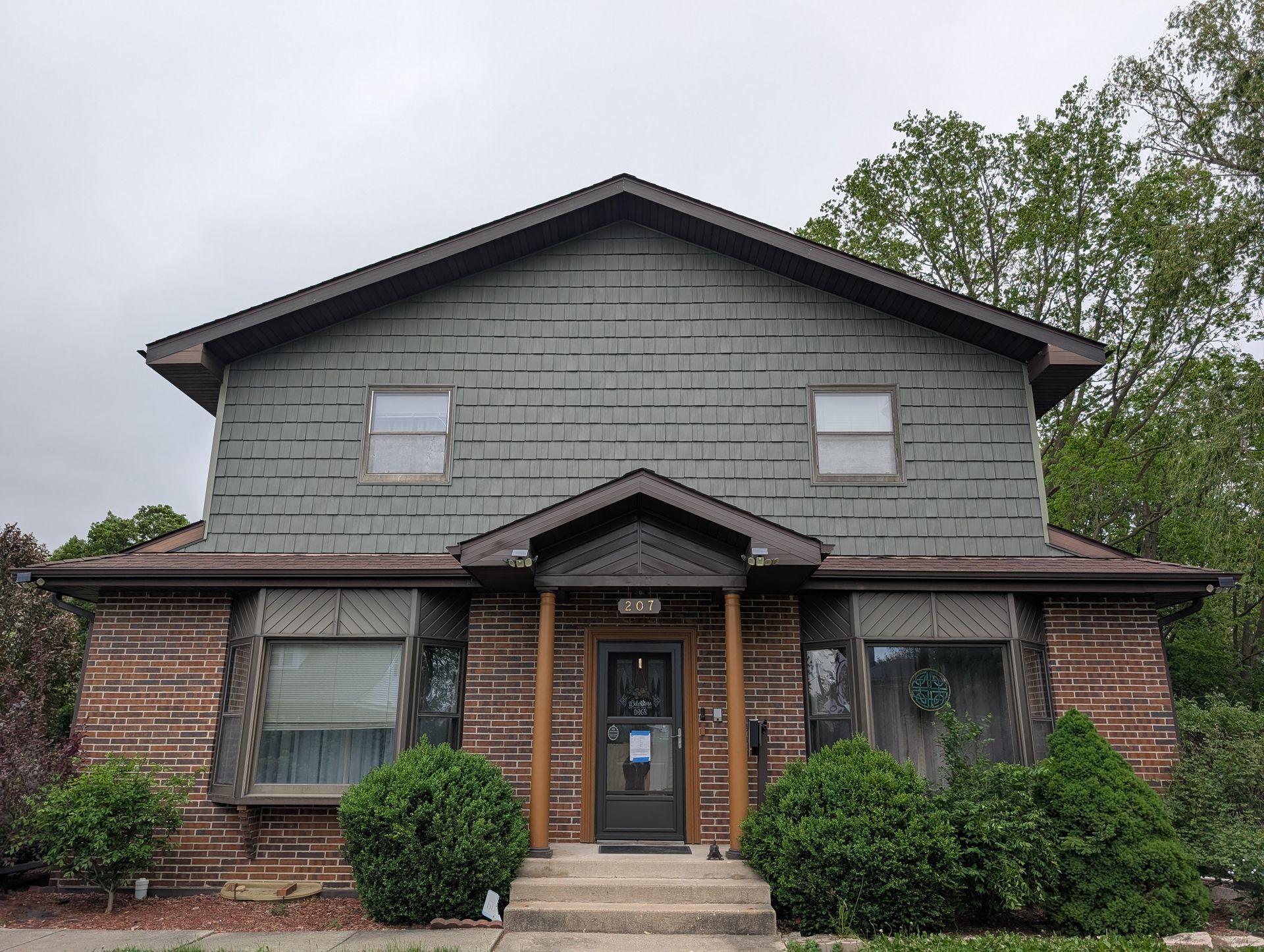 Two-story house with green siding above brick. Brown trim and roof. Bushes in front.