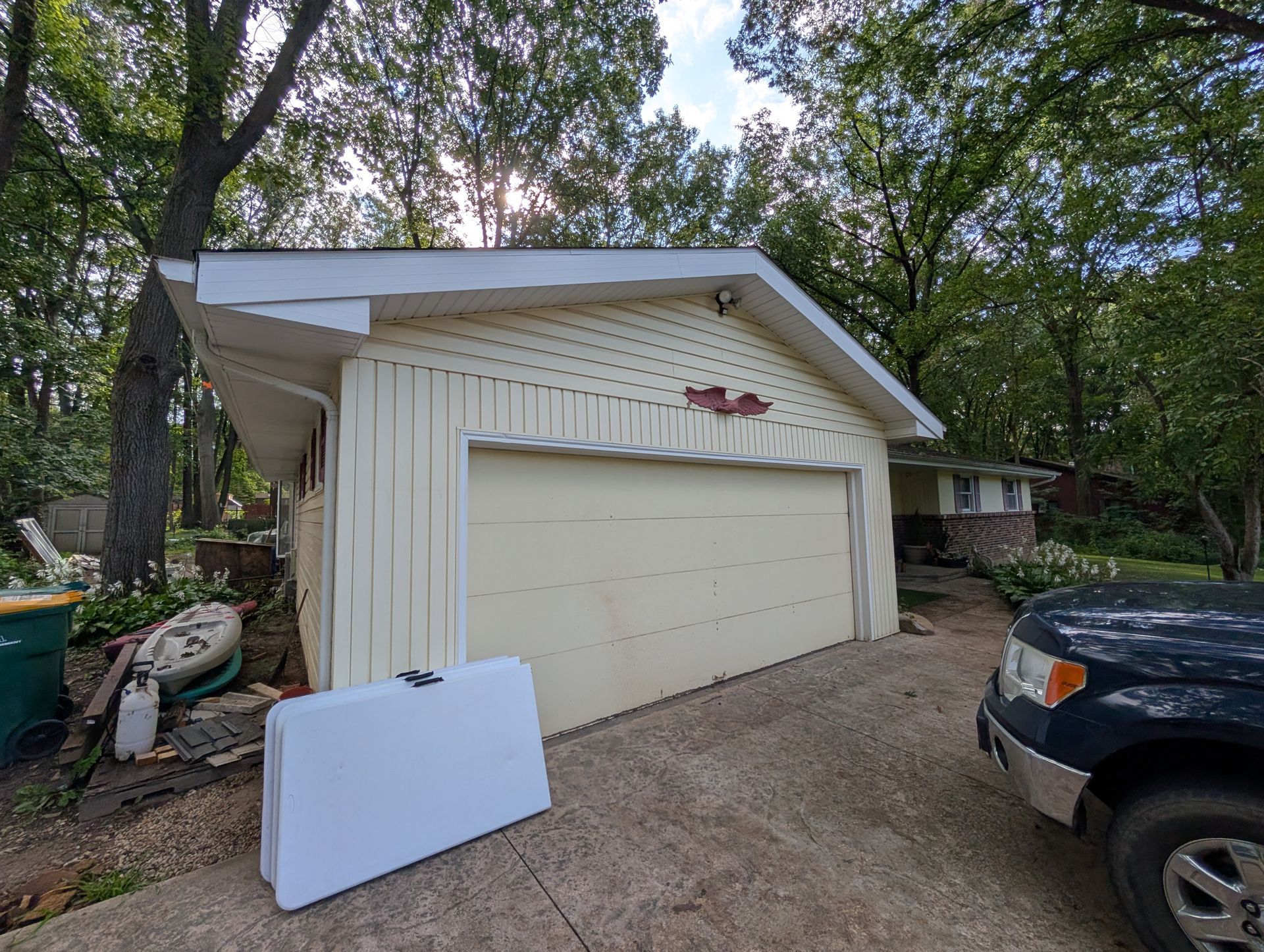 Garage with light yellow siding, closed door, and white table outside on a concrete driveway; car parked next to it.