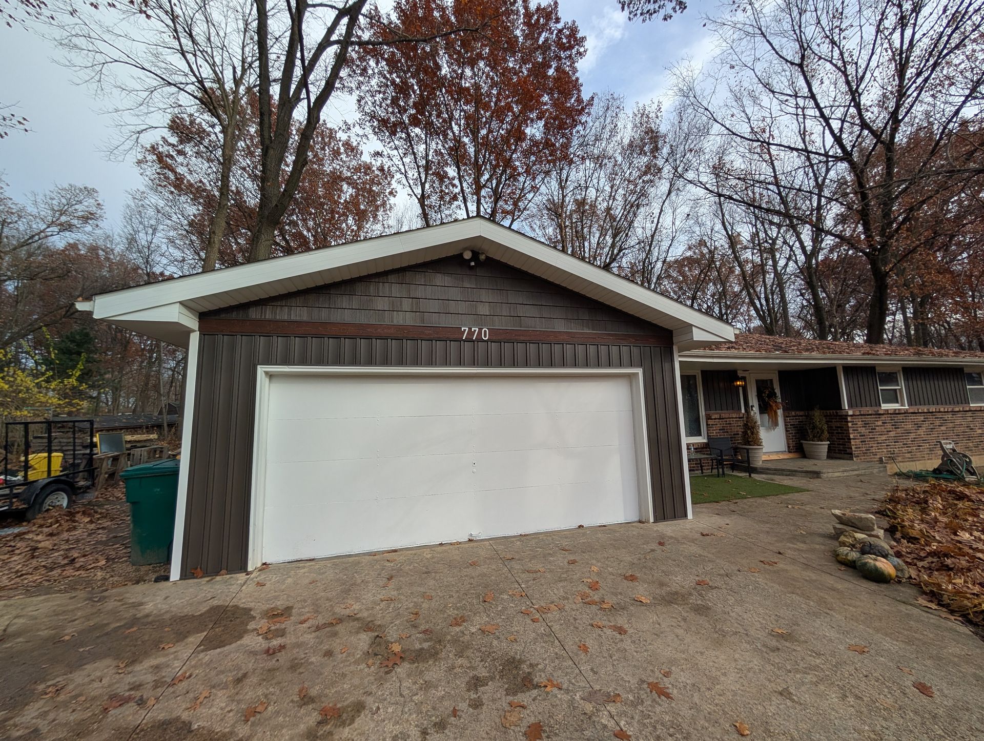 Garage with white door, gray siding, and brown roof. House is to the right with leaves on the ground.