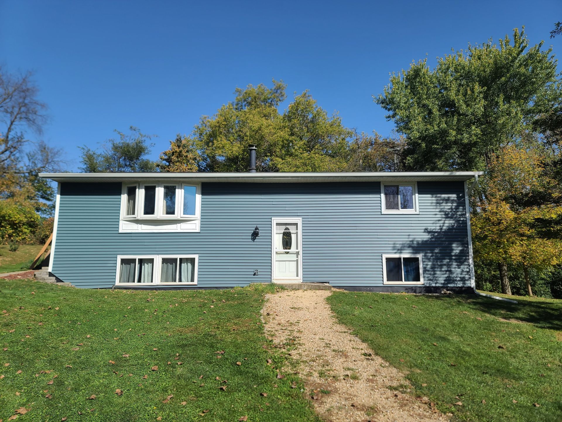 Blue-sided house with white trim, door, and windows, set on a grassy hill, a gravel path leading to the entrance.