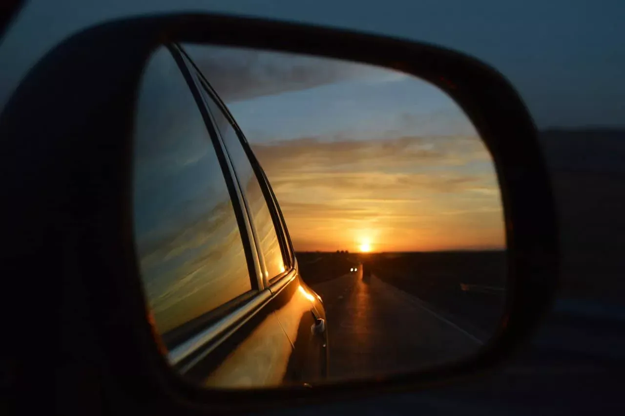 Sunset reflected in car's side mirror, showing the road ahead and the setting sun's orange glow.