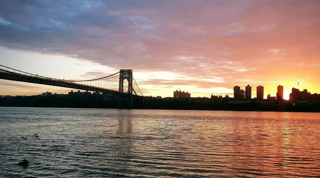 Sunset over a river with a suspension bridge and city skyline silhouetted against the colorful sky.