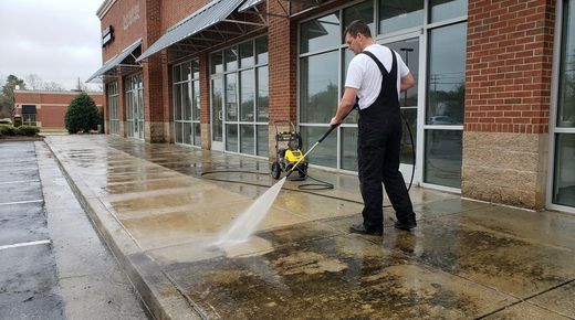 Man pressure washes sidewalk in front of brick storefront using yellow black machine conveying cleanliness and maintenance