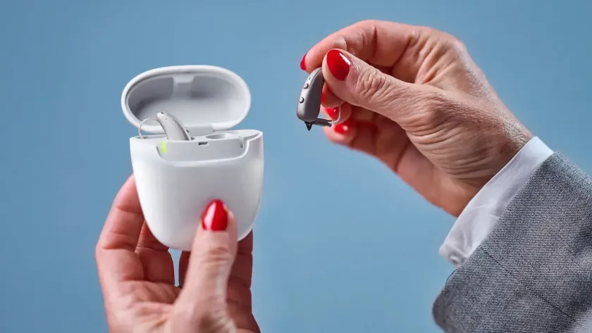 Hands holding a white open charging case and a black earbud against a blue background