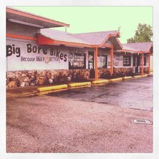 Big Bore Bikes storefront with covered walkway, stone base, and wet parking lot on an overcast day