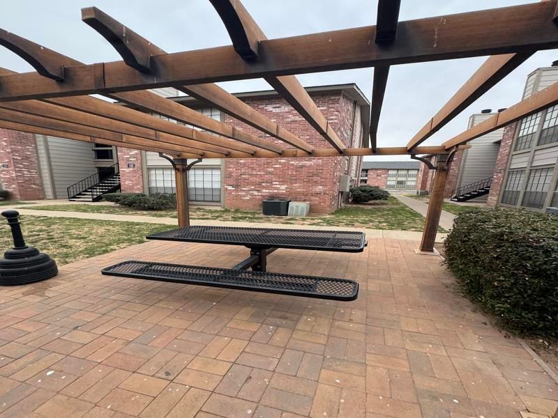 A picnic table under a pergola in front of a brick building.