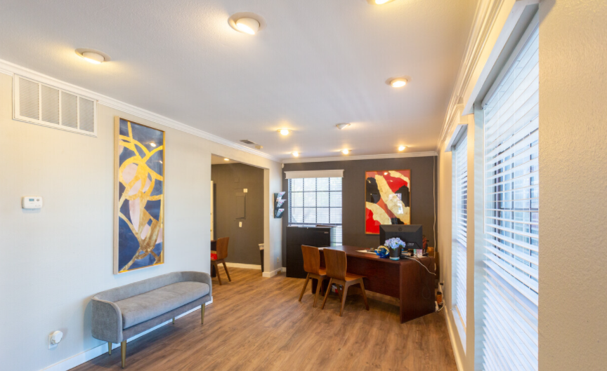 A living room with a bench , desk , chairs and paintings on the wall.