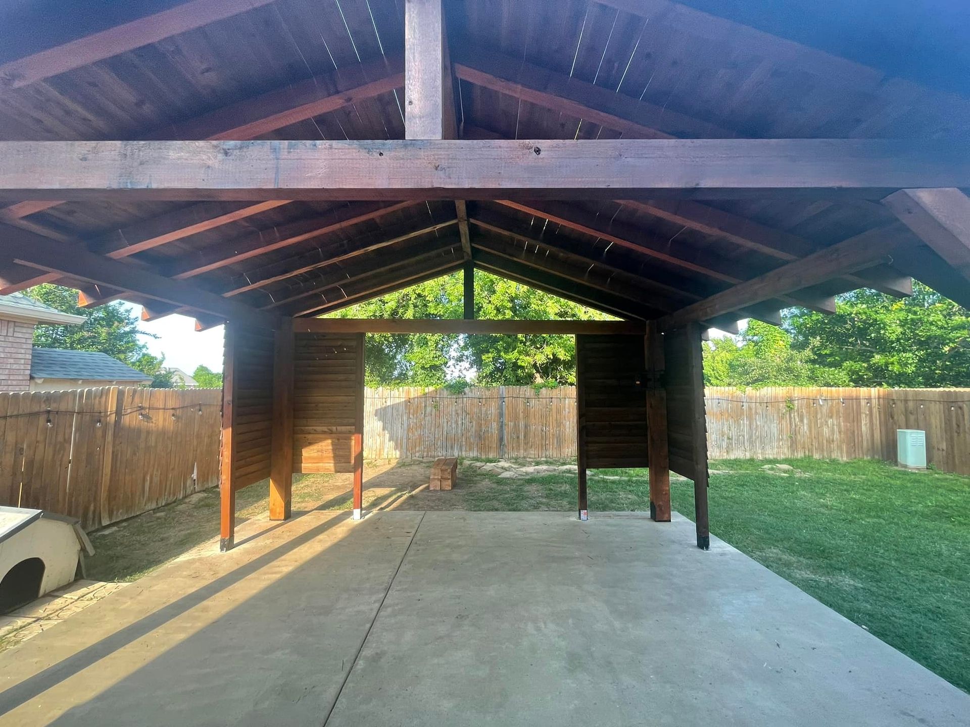 A covered area with a wooden roof and a fence in the background.