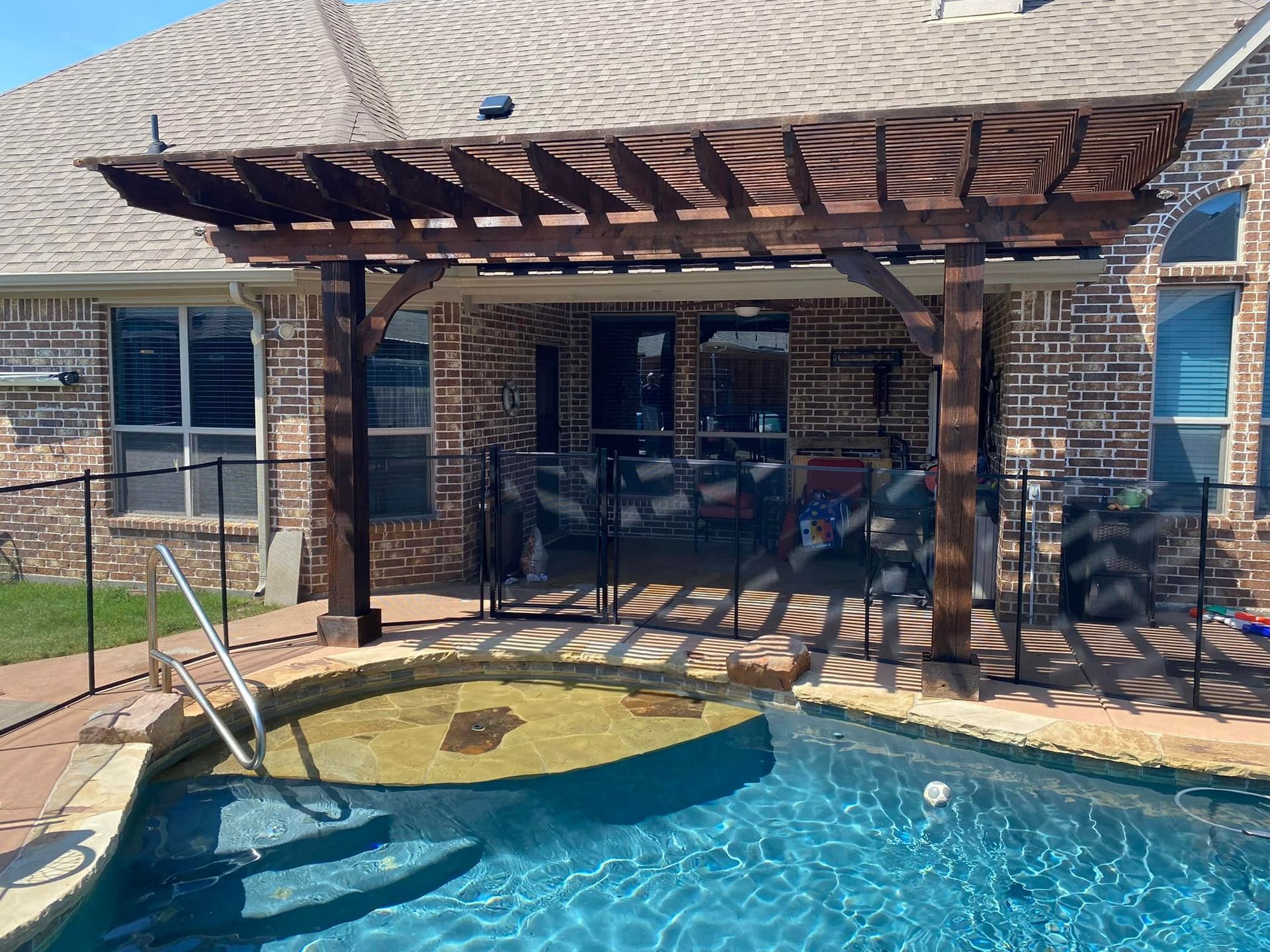 A wooden pergola over a swimming pool in front of a brick house.