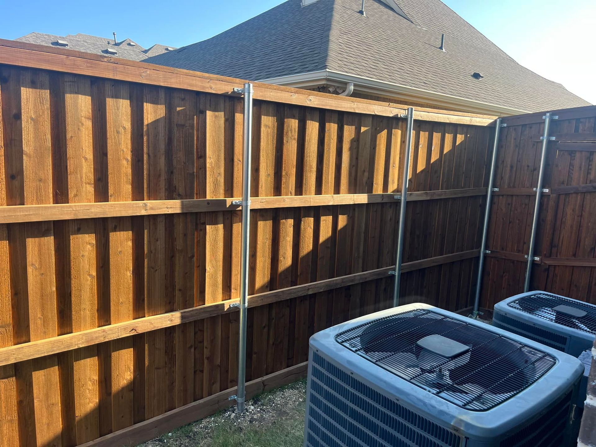 Two air conditioners are sitting next to a wooden fence.