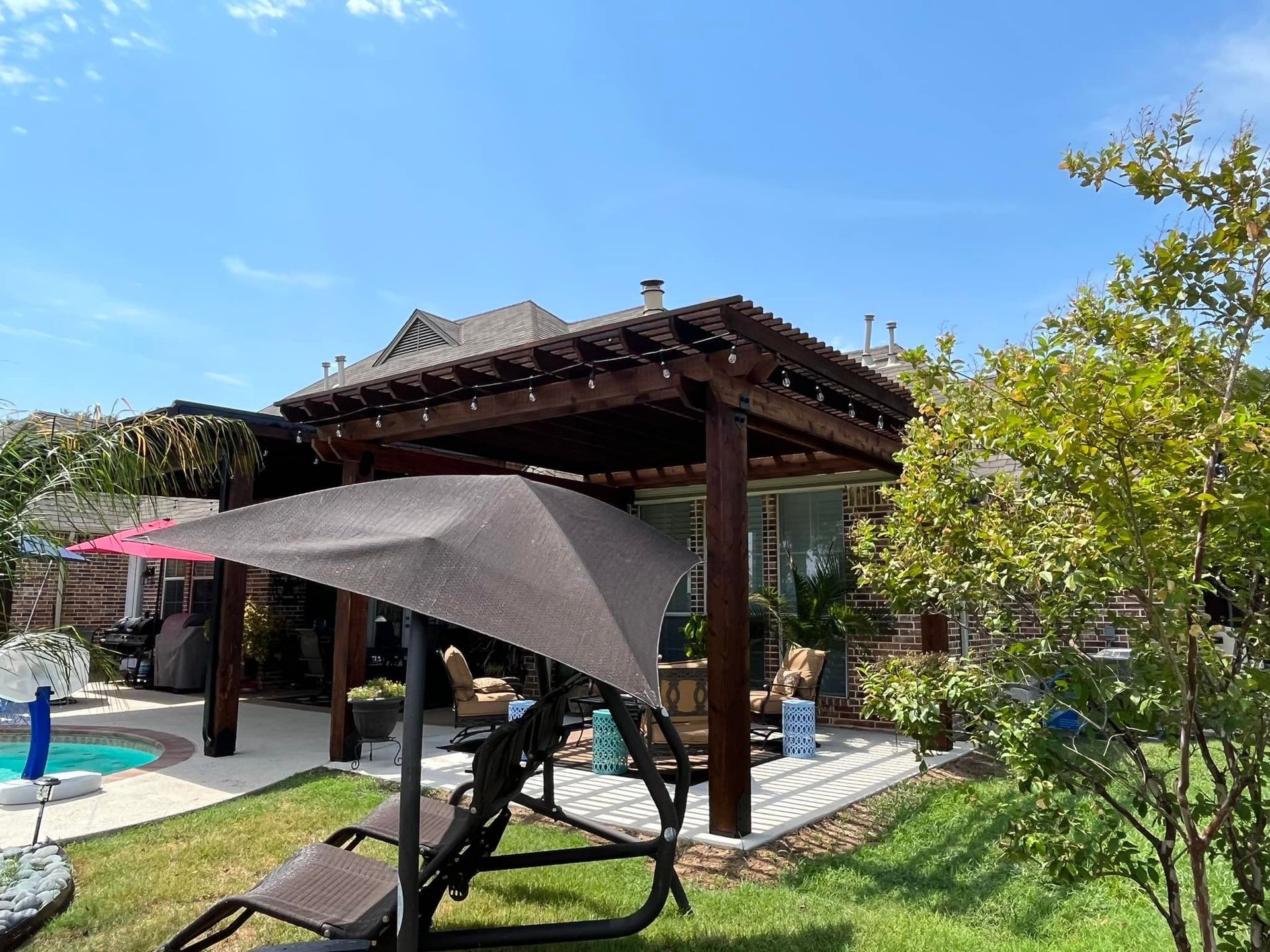 A wooden gazebo with a swing and umbrella in a backyard.