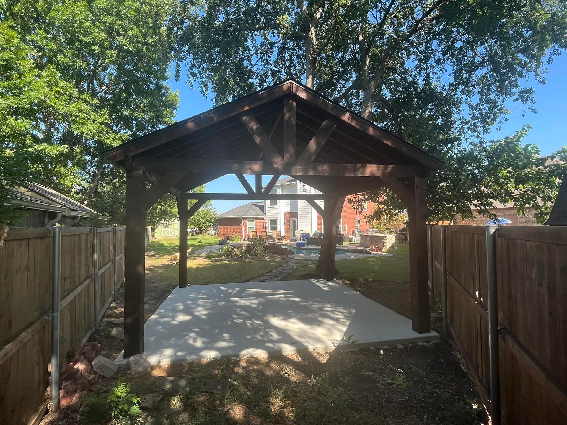 A wooden gazebo in the backyard of a house surrounded by trees and a wooden fence.
