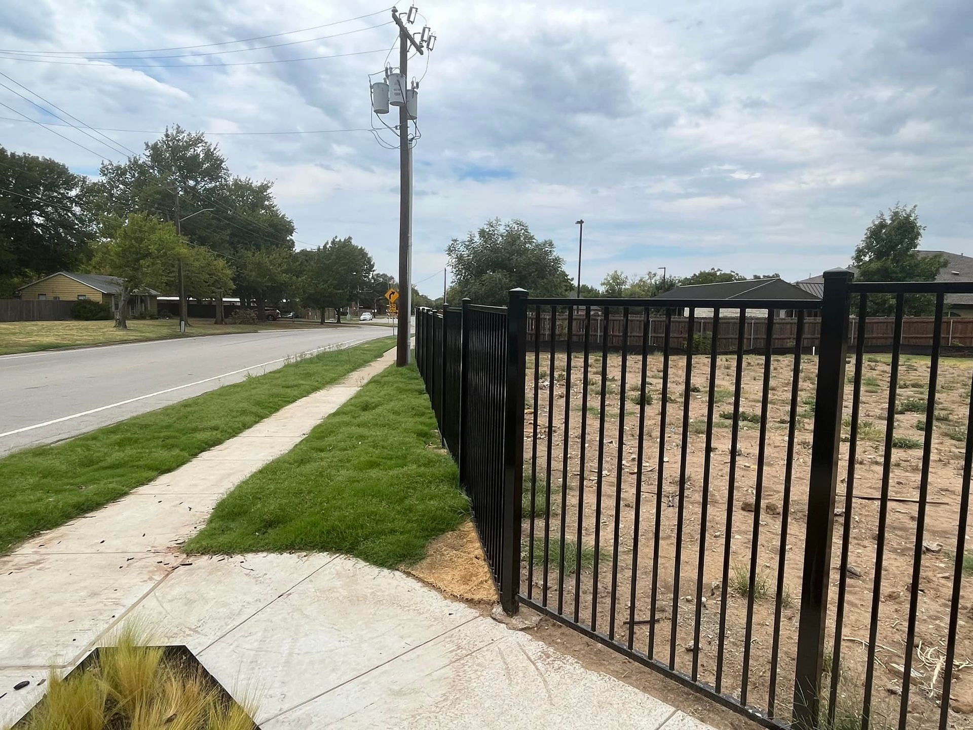 A black fence surrounds a grassy field next to a sidewalk.