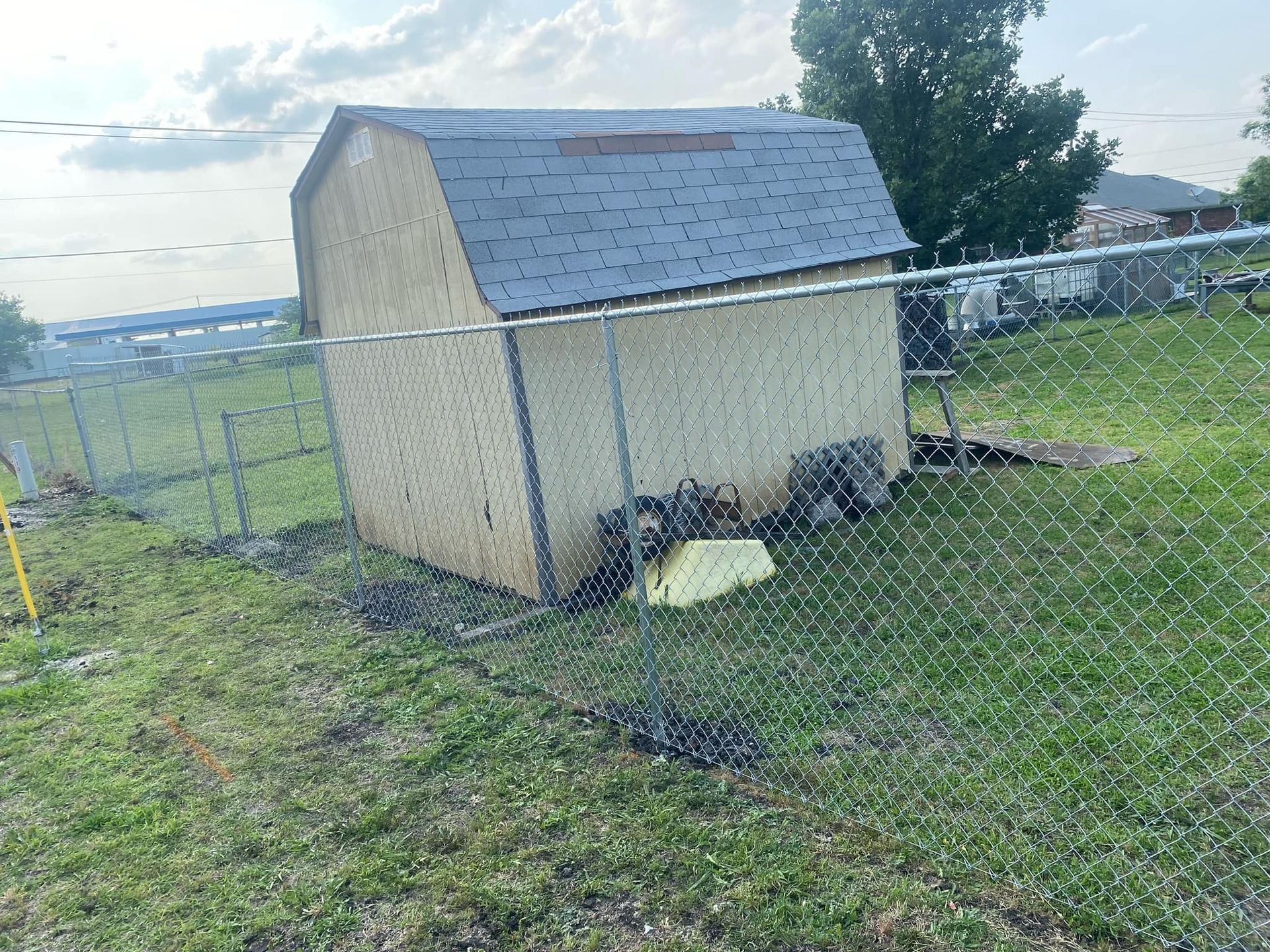 A small shed is sitting in the middle of a grassy field next to a chain link fence.