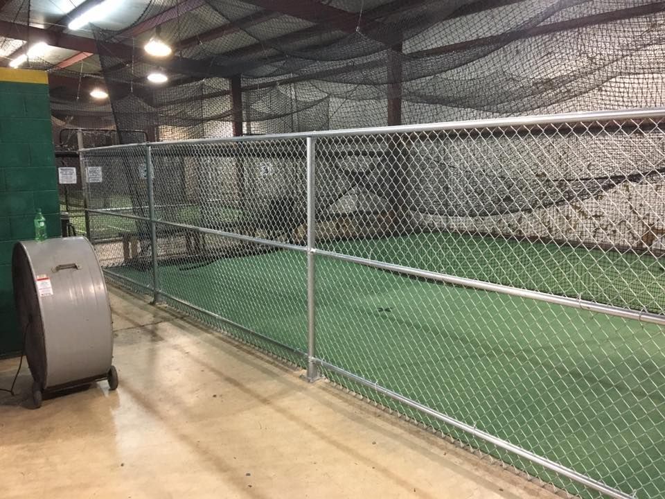 A chain link fence surrounds a baseball field in a warehouse.
