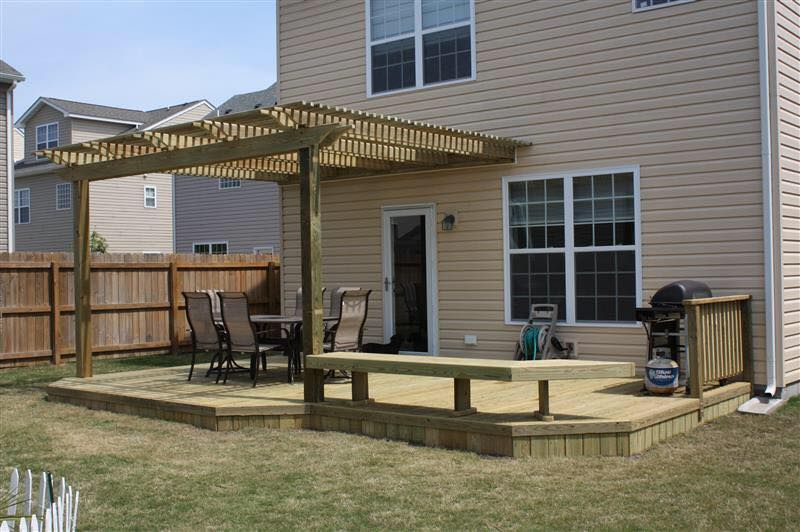 A wooden deck with a pergola and a table and chairs in front of a house.
