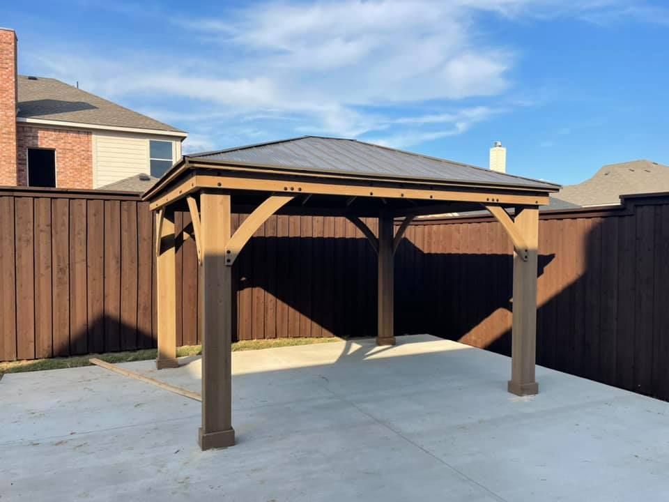 A wooden gazebo with a metal roof is in the backyard of a house.