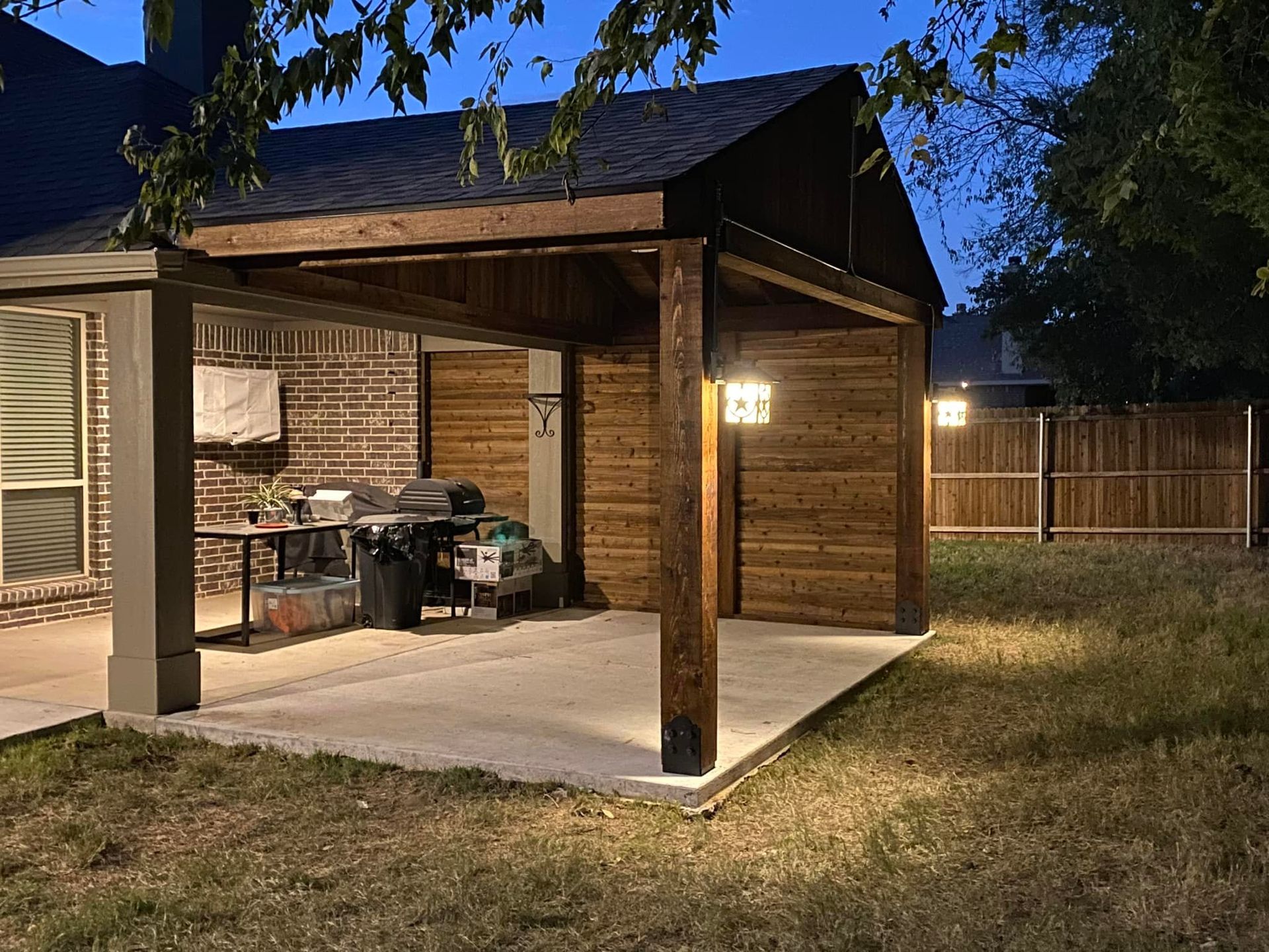 A covered patio with a grill and a fence in the backyard of a house.