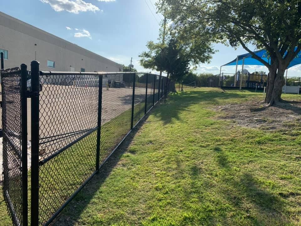 A chain link fence surrounds a lush green field with a playground in the background.