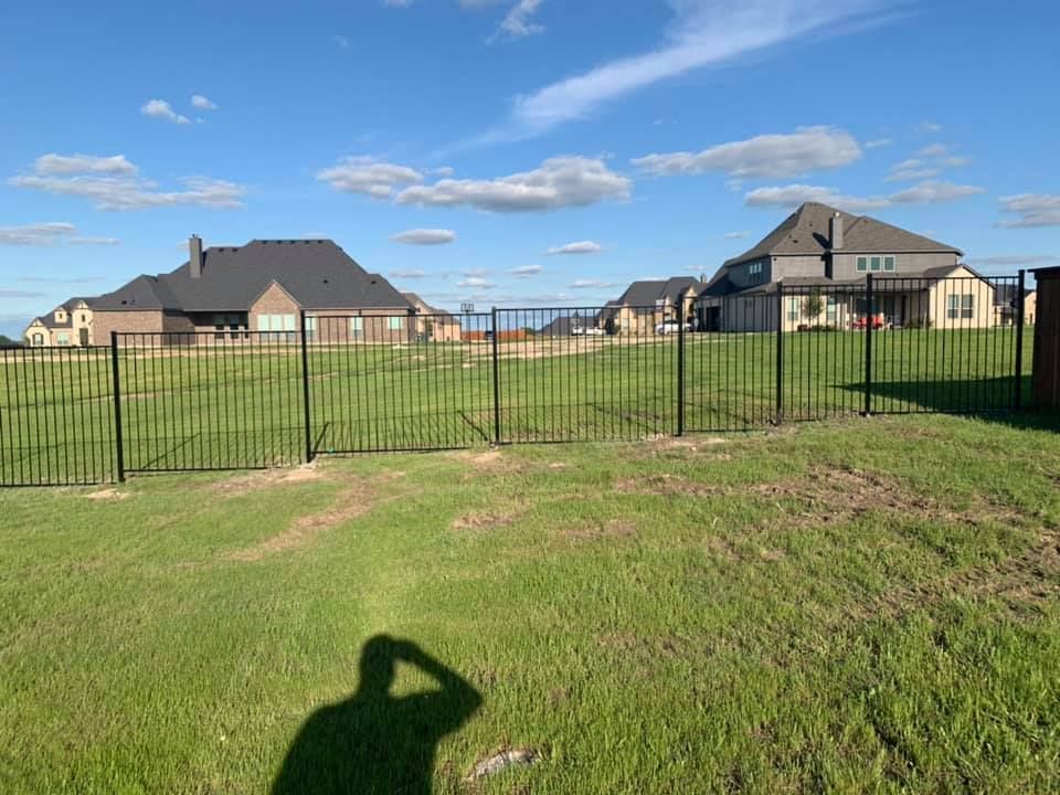 A shadow of a person is cast on the grass in front of a fence.