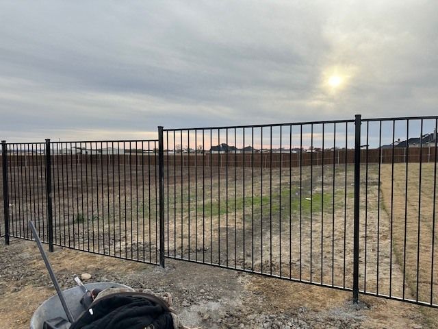 A black fence surrounds a field with the sun shining through the clouds