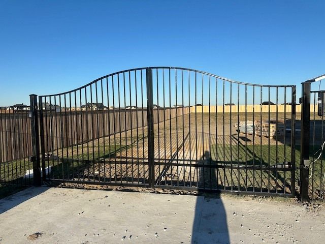 A metal gate is open to a field with a fence in the background.