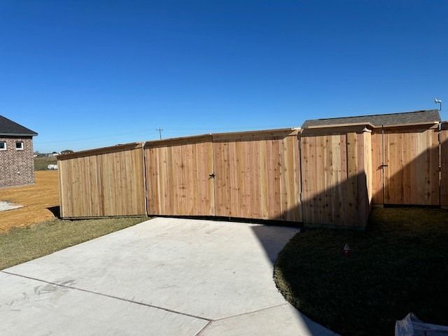 A wooden fence surrounds a driveway with a brick building in the background.