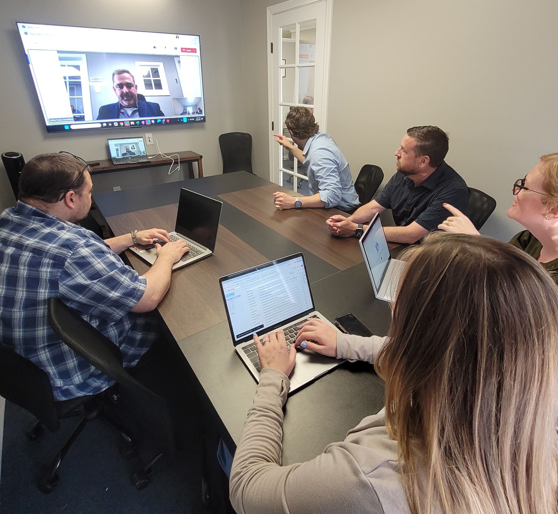 Five people sit at a conference table with laptops during a remote meeting viewed on a large wall-mounted screen.