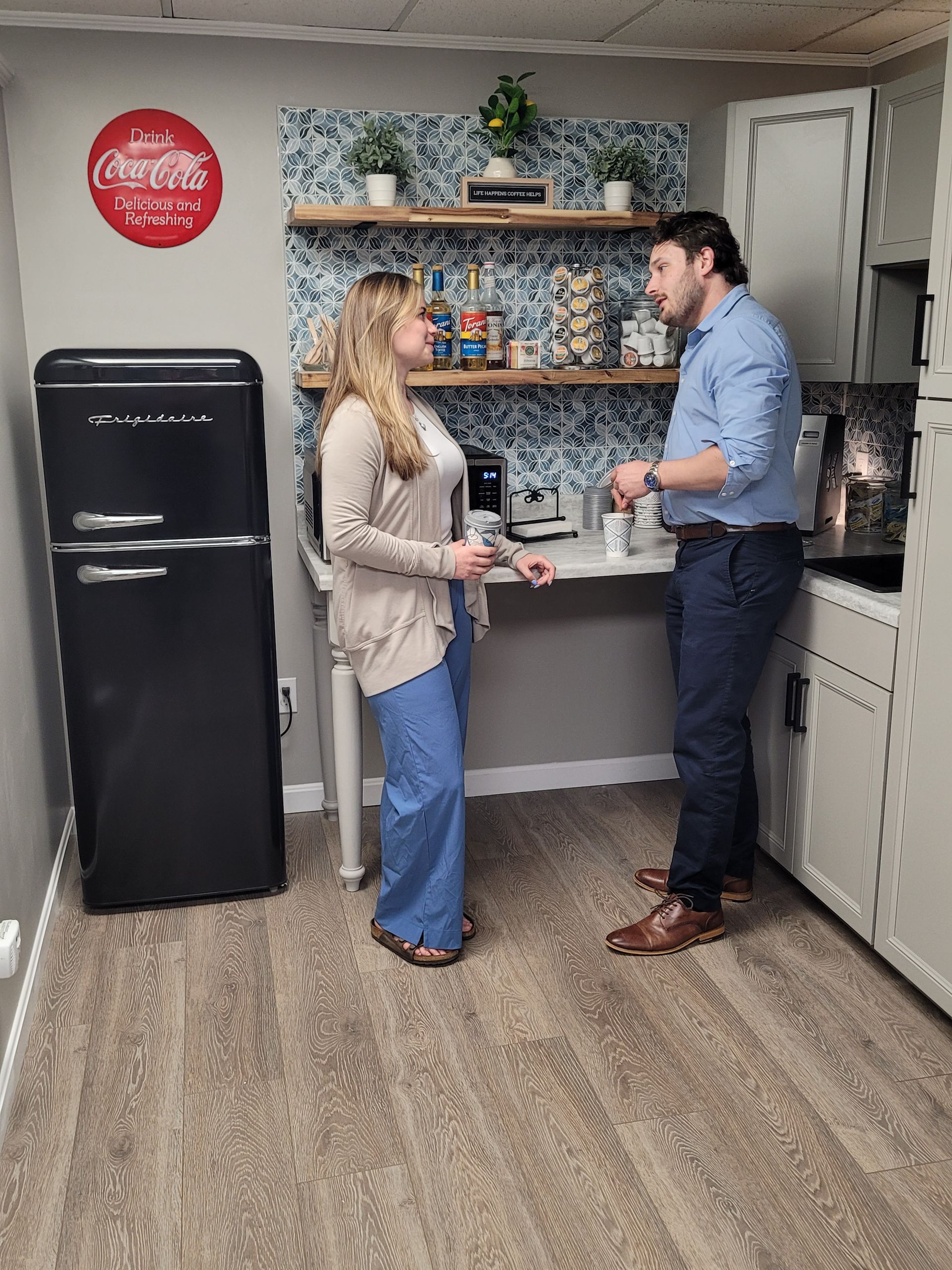 A person in a beige cardigan and blue pants stands in a kitchen, talking to a person in a blue shirt and dark trousers.